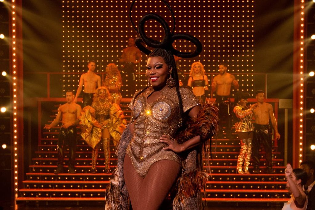 화려한 의상과 스포트라이트를 배경으로 무대에서 커다란 헤드피스를 입은 금색 의상을 입은 댄서.