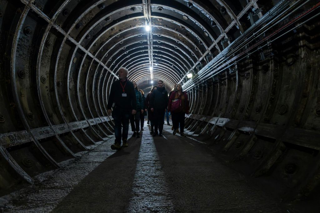 곡선형 금속 벽과 머리 위 조명이 있는 어두운 지하 터널을 걷고 있는 한 무리의 사람들.