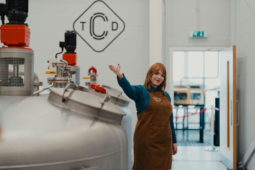 갈색 앞치마를 입은 여성이 증류소에 서서 미소를 짓고 제스처를 취하고 있으며, 장비와 로고가 배경에 보입니다.