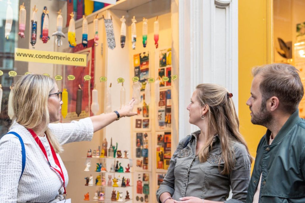 두 사람이 알록달록한 콘돔과 다양한 소품이 진열된 상점을 바라보고 있습니다. 다른 사람이 디스플레이를 향해 제스처를 취합니다.