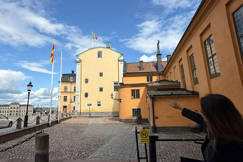 자갈 바닥, 노란색 건물, 깃발, 푸른 하늘 아래 오른쪽에서 몸짓을 하는 사람이 있는 안뜰.