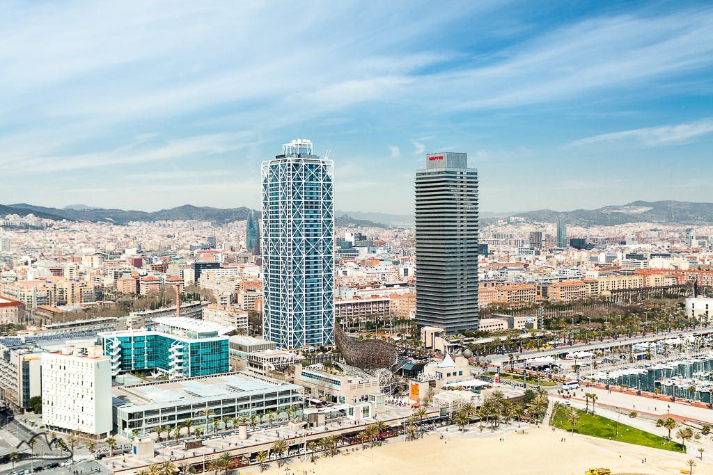 두 개의 높은 고층 건물, 주변 건물, 전경의 모래 해변, 푸른 하늘이 있는 도시 풍경.