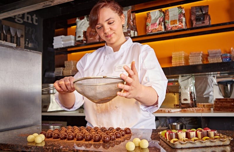 흰색 코트를 입은 셰프가 주방에서 초콜릿 송로버섯 위에 코코아 가루를 뿌립니다. 재료가 담긴 선반이 배경입니다.