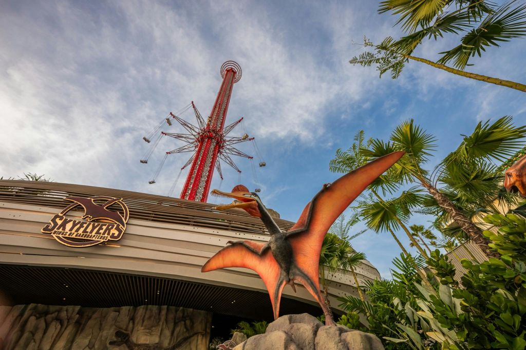 놀이공원 입구 앞에는 주황색 날개를 가진 날아다니는 공룡 동상이 있으며, 배경에는 빨간색의 키가 보입니다.