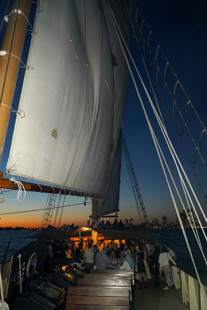 저녁에는 대형 돛이 달린 범선이 정박하거나 유람하며, 선상에는 사람들이 타고 있고 배경에는 도시의 스카이라인이 보입니다.