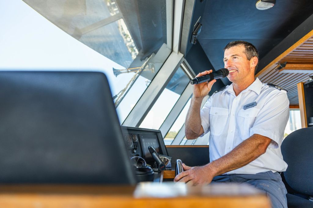 제복을 입은 남성이 커다란 창문이 있는 보트 옆에서 마이크를 들고 이야기하고 있습니다.