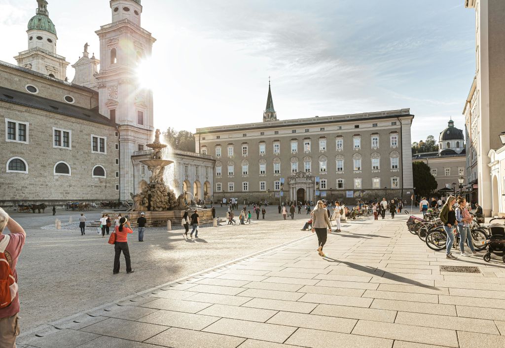분수와 커다란 건물, 교회 타워를 배경으로 햇살이 내리쬐는 광장 주변을 사람들이 거닐고 있습니다.