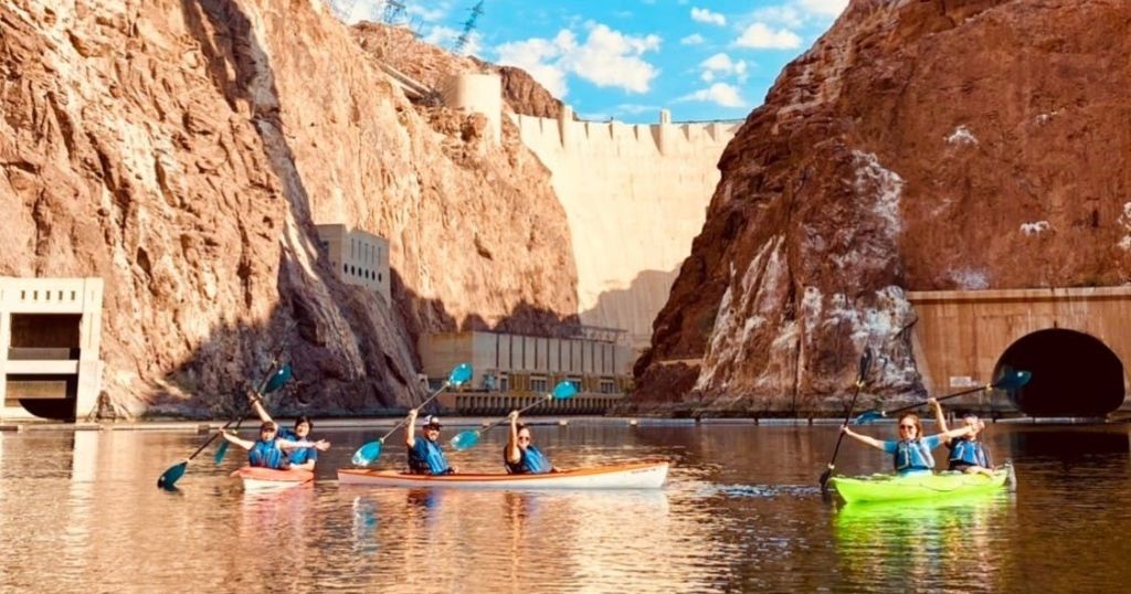 흩어진 구름과 함께 푸른 하늘 아래 커다란 댐과 바위 절벽 근처에서 잔잔한 물 위에서 카약을 타는 한 무리의 사람들.
