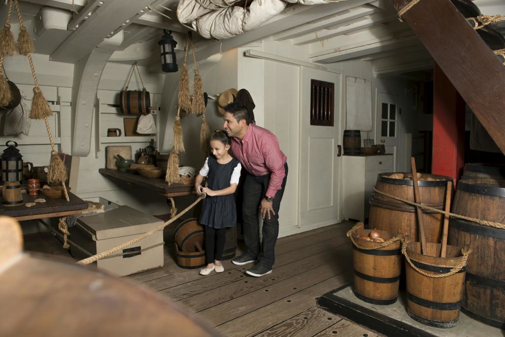 소박한 배의 오두막 안에 밧줄, 배럴, 해상 장식 근처에 서 있는 한 남자와 소녀가 소녀의 어깨에 손을 얹고 몸을 앞으로 숙이고 있습니다.