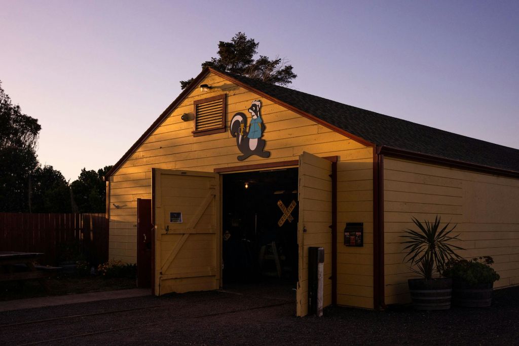 황혼에 열린 문이 있는 노란색 헛간으로, 외관에 타이어가 달린 만화 스컹크가 특징입니다. 내부에는 도구와 철도 건널목 표지판이 보입니다.