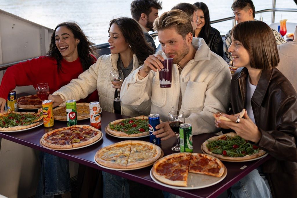 야외 데크의 테이블에서 3명이 다양한 피자와 음료를 즐기고 있으며, 배경에는 물줄기가 보입니다.