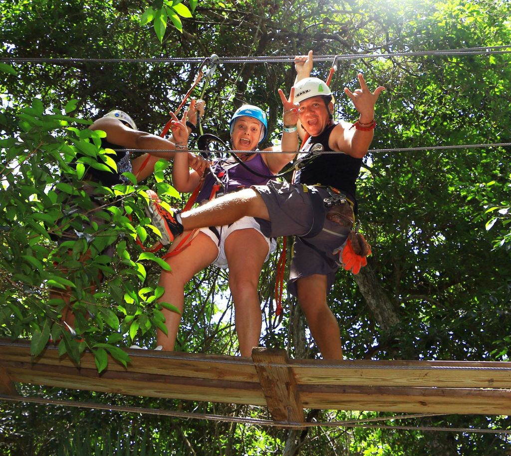 헬멧과 마구를 쓴 세 사람이 나무 사이에 있는 나무 플랫폼에 서서 열정적인 손짓을 하고 있습니다.