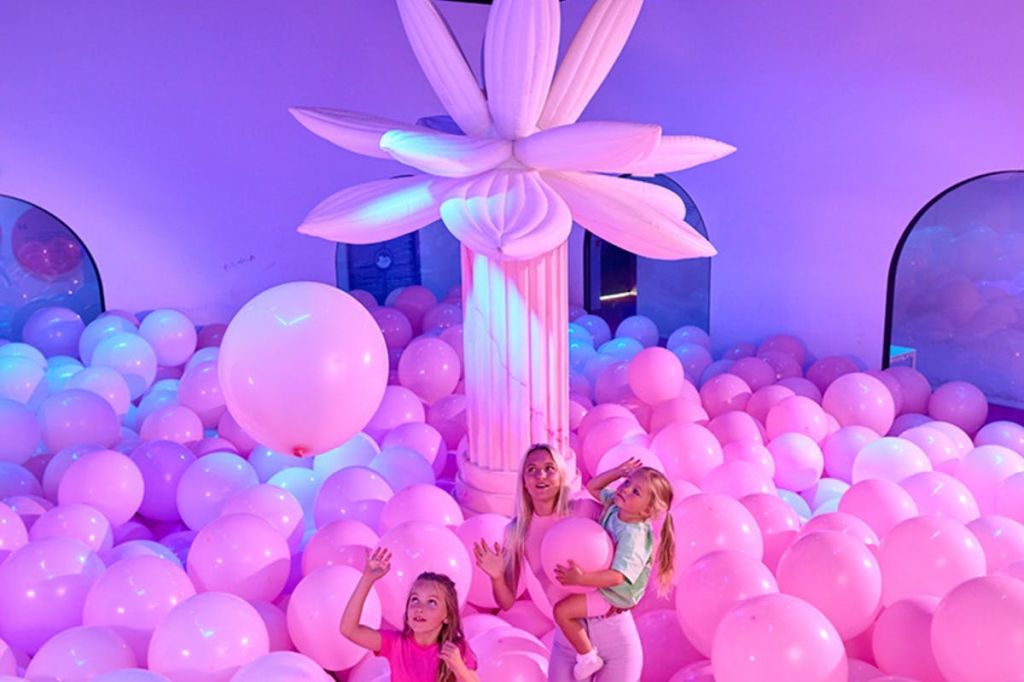 분홍색과 보라색 풍선이 가득한 방에서 한 여성과 두 자녀가 기둥에 부착된 커다란 팽창식 꽃 옆에서 놀고 있습니다.
