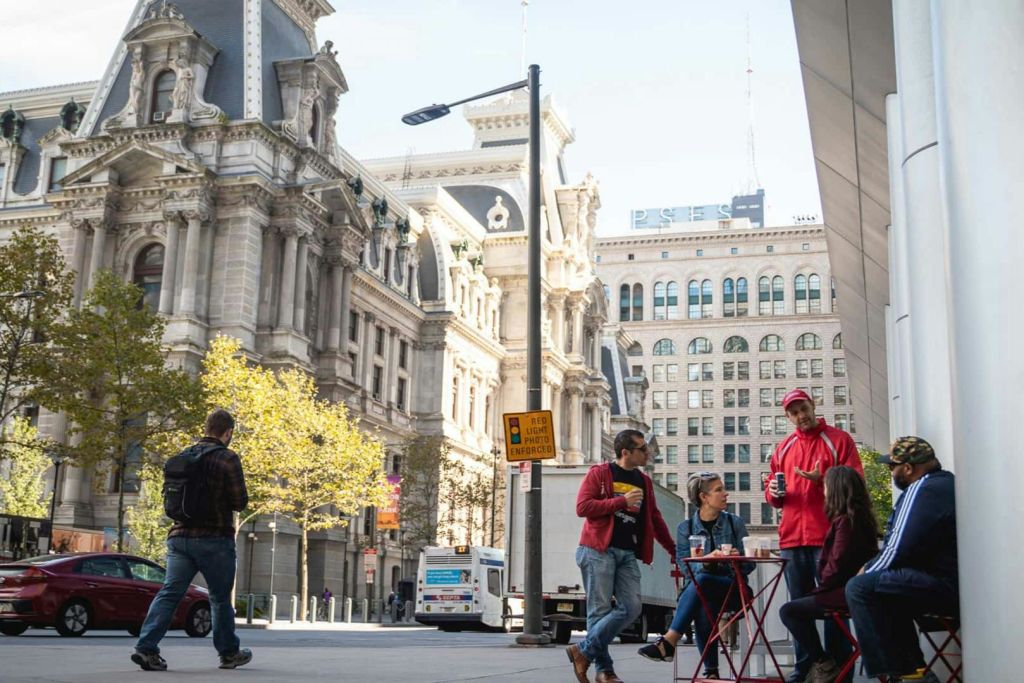 유서 깊은 건물과 주차된 버스를 배경으로 도시 거리의 작은 바깥 테이블에 네 사람이 모여듭니다.