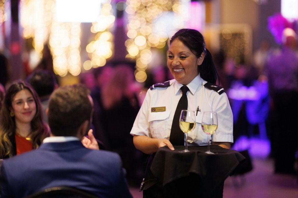 유니폼을 입고 와인 한 잔과 함께 트레이를 들고 있는 서버는 따뜻하게 빛나는 축제 분위기에 앉아 커플을 맞이합니다.