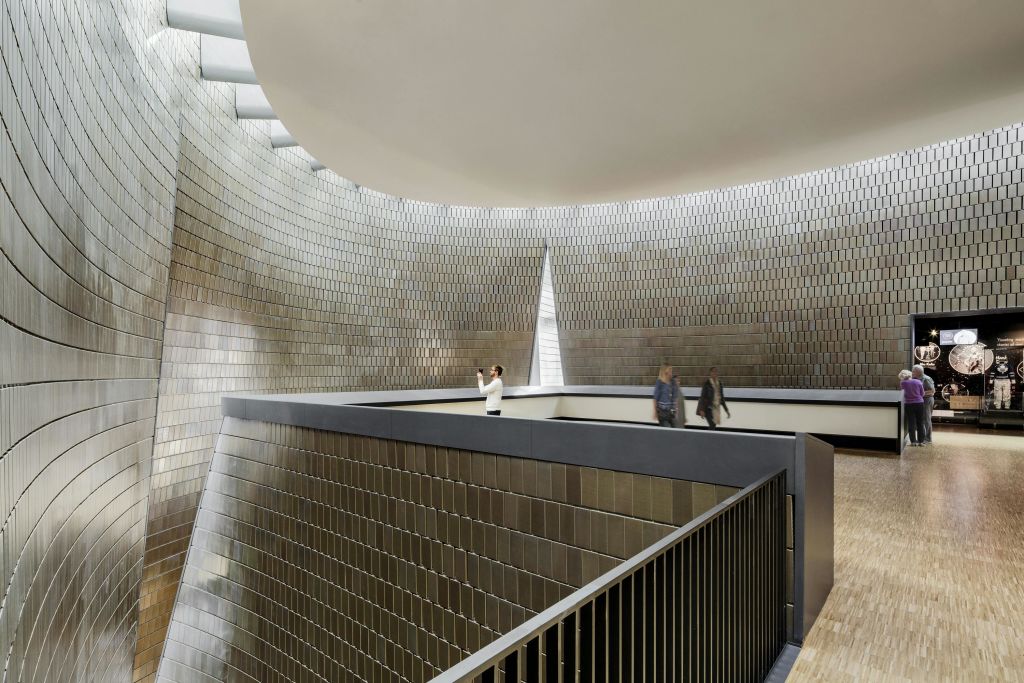 금속 타일로 마감된 벽과 높고 곡선형의 천장이 있는 현대적이고 넓은 건물에서 세 사람이 발코니를 거닐고 있습니다.