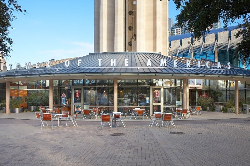 "OF THE AMERIC" 이라고 표시된 유리 벽 건물 앞에 빈 테이블과 의자가 있는 야외 카페. “뒤에 우뚝 솟은 구조물.