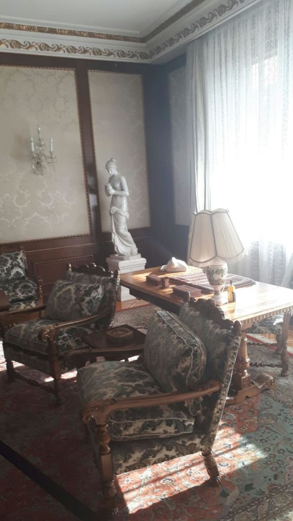 앤티크 가구, 커다란 책상, 화려한 의자, 조각상, 램프, 레이스 커튼을 통과하는 조명 필터링이 있는 객실입니다.