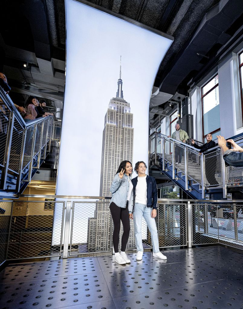 현대적인 전망대 안에 있는 엠파이어 스테이트 빌딩의 대형 디스플레이 앞에서 두 사람이 사진을 찍기 위해 포즈를 취하고 있습니다.