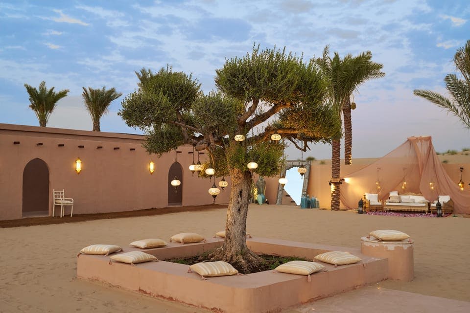 등불로 장식된 나무가 있는 마당으로, 모래사장에 베개로 둘러싸여 있으며, 야자수와 건물을 배경으로 하고 있습니다.