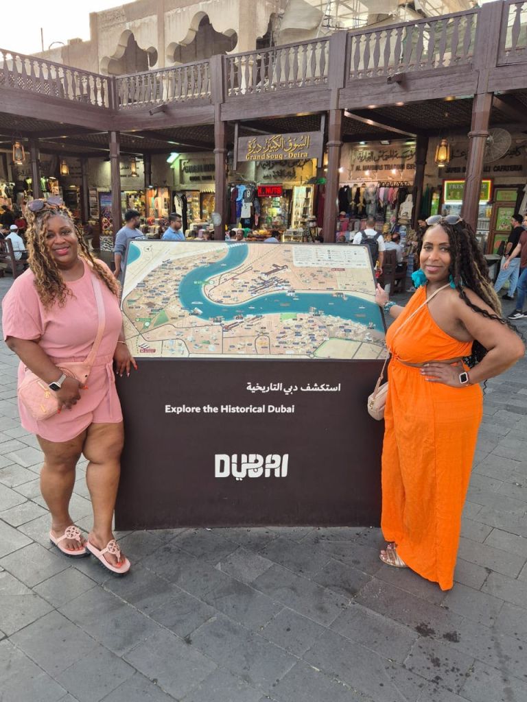 두 명의 여성이 상점과 사람들을 배경으로 분주한 시장에서 역사적인 두바이의 관광 지도 옆에서 포즈를 취하고 있습니다.