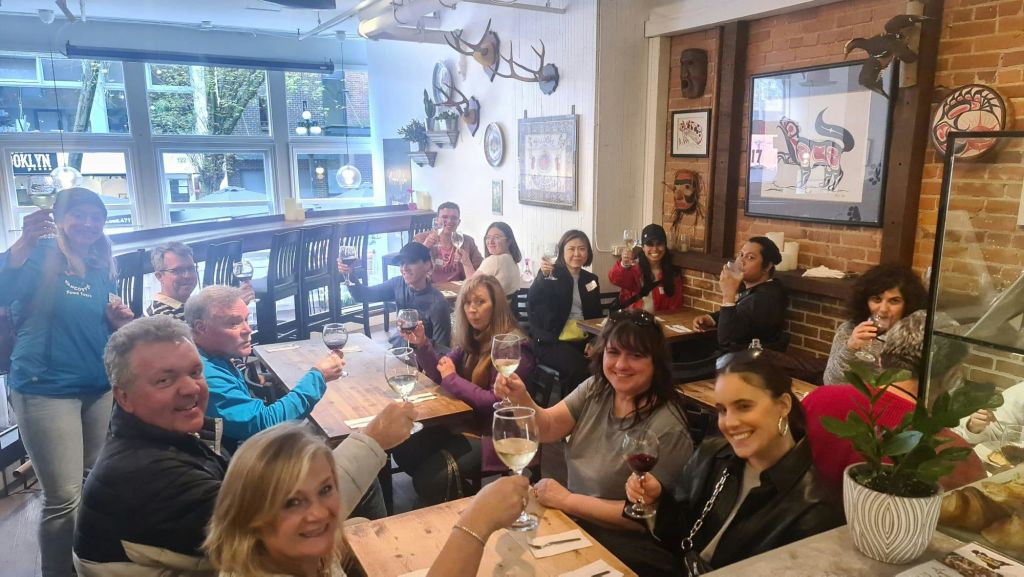 한 무리의 사람들이 소박한 레스토랑의 나무 테이블에 앉아 와인잔을 들고 카메라를 보며 미소를 짓고 있습니다.