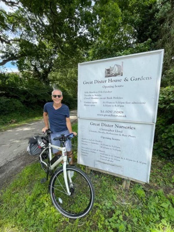 한 사람이 나무로 둘러싸인 '그레이트 딕스터 하우스 & 가든' 과 '그레이트 딕스터 보육원' 표지판 앞에 자전거 옆에 서 있습니다.