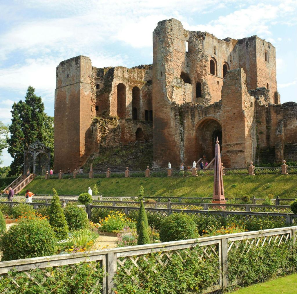 아치형 창문이 있는 부분적으로 폐허가 된 고대 석조 성으로, 잘 손질된 수풀과 꽃이 피는 식물이 있는 정원 뒤에 자리 잡고 있습니다.