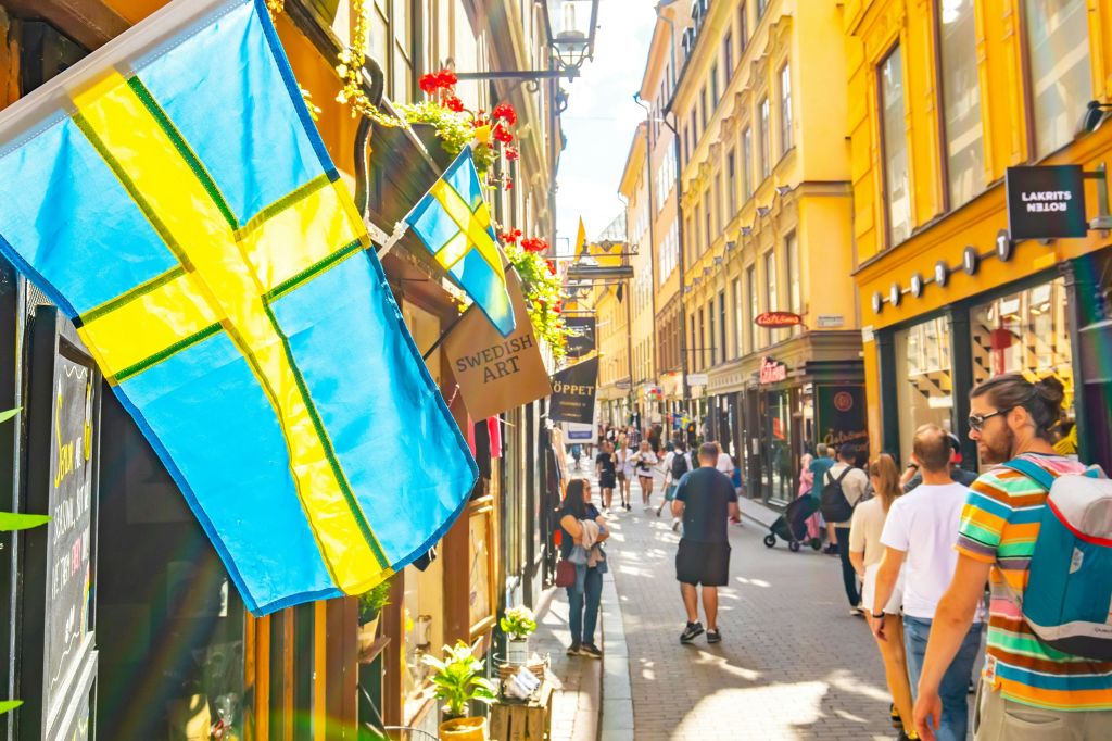 걷는 사람들, 화려한 건물, 스웨덴 국기가 전시된 유럽 도시의 활기찬 보행자 거리입니다.