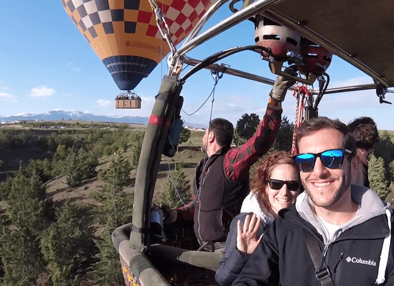 세 사람이 열기구 바구니 안에서 미소를 짓고 손을 흔들고 있으며, 다른 풍선이 배경에 보입니다.