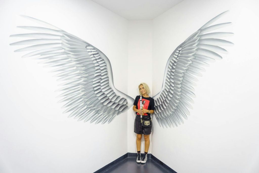 한 사람이 흰 방 구석에 서서 벽에 페인트칠된 천사 날개 사이에서 포즈를 취하고 있습니다. 그들은 음료를 들고 캐주얼한 옷을 입습니다.