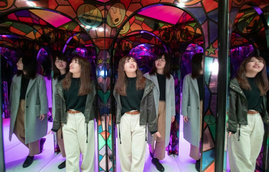 형형색색의 거울처럼 빛나는 터널에 서서 여러 개의 반사를 만들어내는 여성. 밝은 바지와 어두운 상의, 코트를 입고 있습니다.