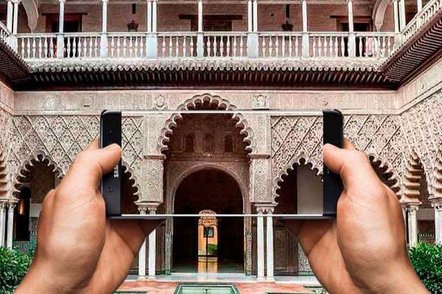 정교한 조각이 돋보이는 화려한 석조 아치형 통로와 아치 너머로 보이는 안뜰을 담은 스마트폰을 들고 있는 사람.