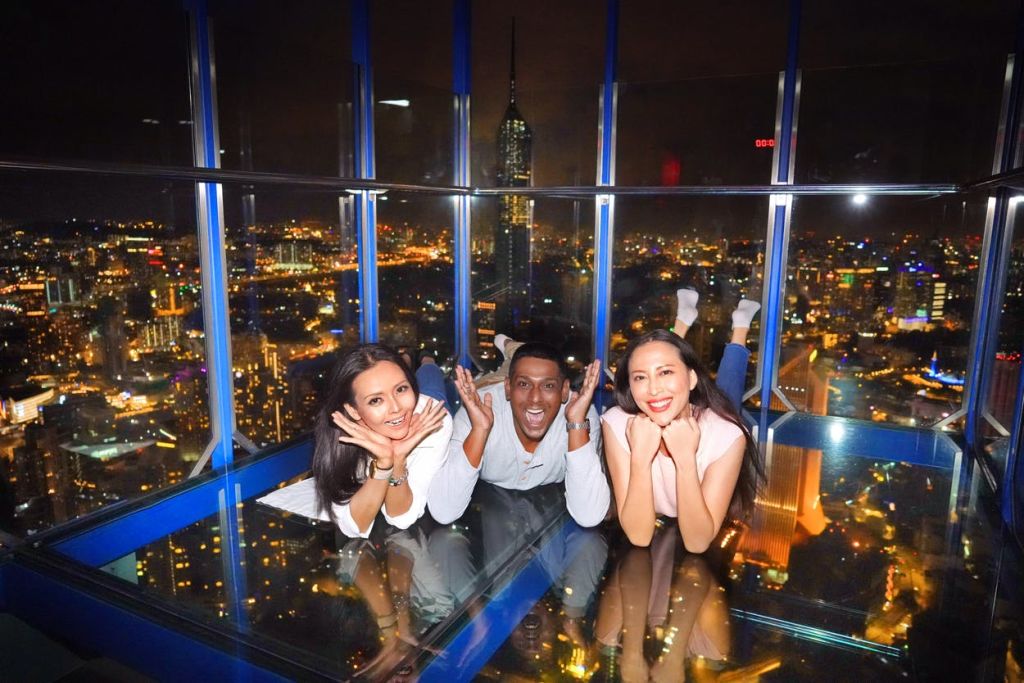 세 사람이 유리 바닥에 누워 포즈를 취하고 미소를 짓고 있으며, 밤에는 도시 풍경이 배경으로 펼쳐집니다.