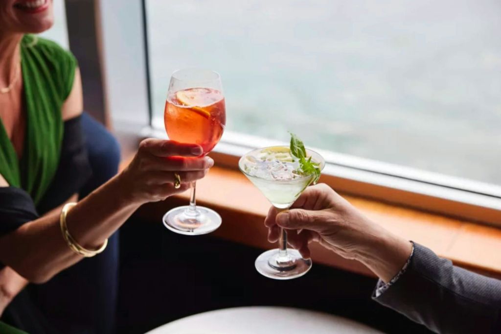 창문 근처에서 칵테일을 들고 있는 두 손, 하나는 오렌지 조각이 들어간 빨간색 음료, 다른 하나는 장식이 들어간 초록색 음료입니다.