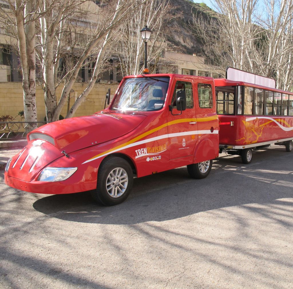 잎이 없는 나무가 늘어선 거리에 선실과 탁 트인 객차가 있는 밝은 빨간색 관광 열차입니다.