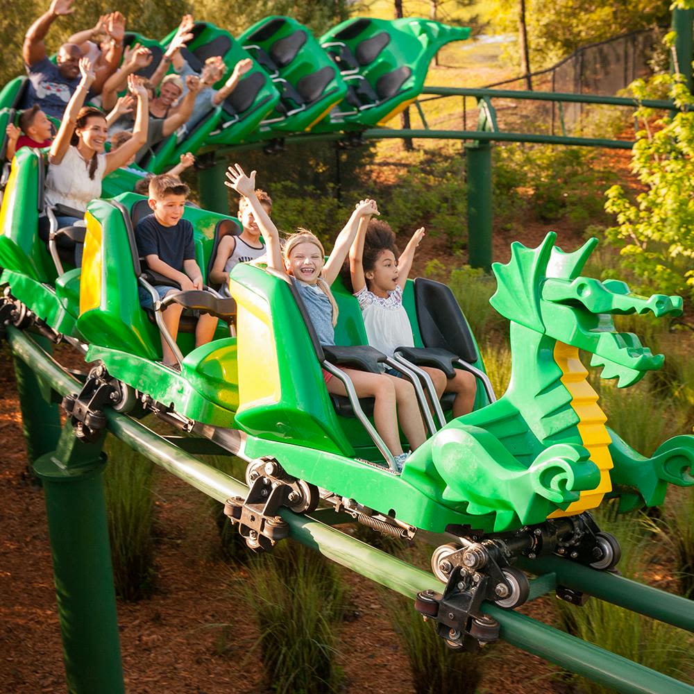 그린 드래곤을 주제로 한 롤러코스터를 타는 어린이들이 나무와 녹지로 둘러싸인 채 미소를 짓고 손을 들고 있습니다.