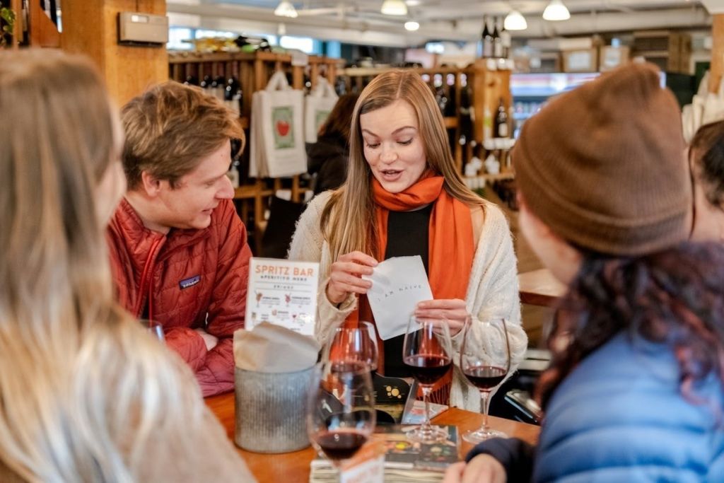 와인 가게의 나무 테이블에 네 명이 앉아 레드 와인 잔과 스프리츠 바를 홍보하는 작은 간판을 들고 앉아 있습니다.