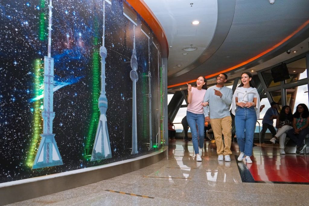 세 사람이 미소를 지으며 근처에 앉아 있는 다른 사람들과 함께 현대적이고 조명이 밝은 방 안에 있는 타워와 건물의 커다란 벽 디스플레이를 가리키고 있습니다.