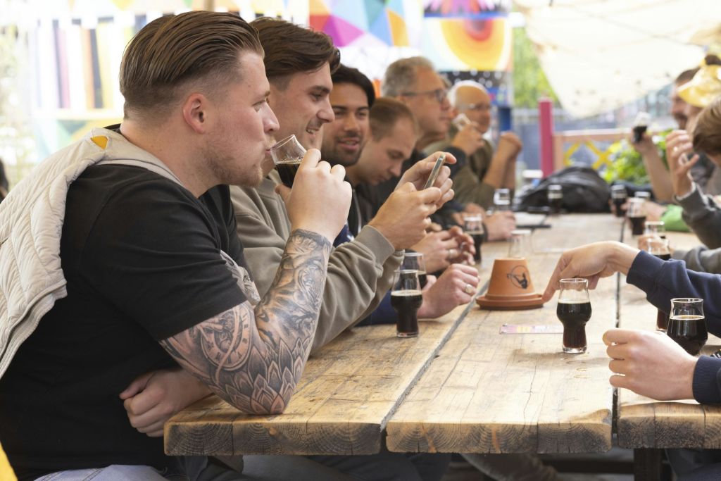 긴 나무 테이블에 앉아 술을 마시며 친목을 도모하는 사람들의 모임. 초점의 남자는 문신을 하고, 일부는 휴대폰을 봅니다. 장식된 배경.