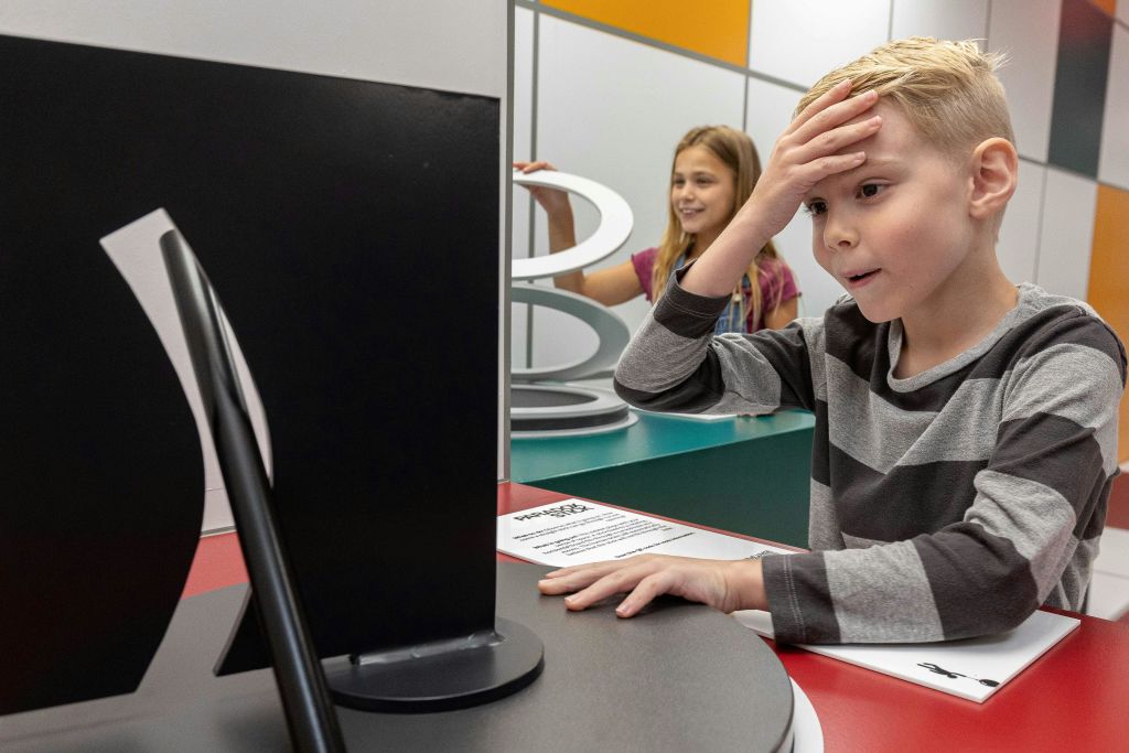 줄무늬 셔츠를 입은 한 소년이 이마를 만지며 어리둥절한 표정을 짓고 있습니다. 한 소녀가 웃으며 배경의 디스플레이와 상호 작용하고 있습니다.