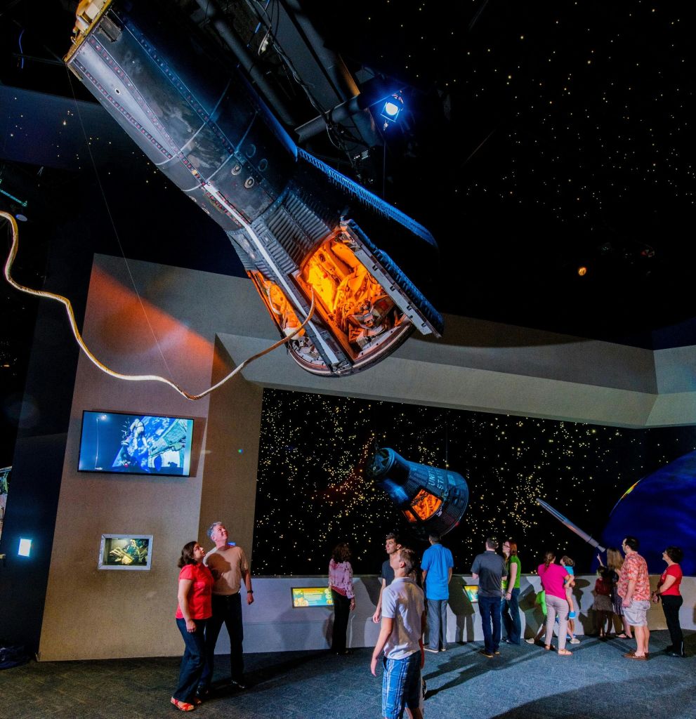 별이 빛나는 전시물과 정보 화면이 있는 희미한 조명의 박물관에서 정지된 우주선을 포함한 우주 전시물을 관람하는 사람들.