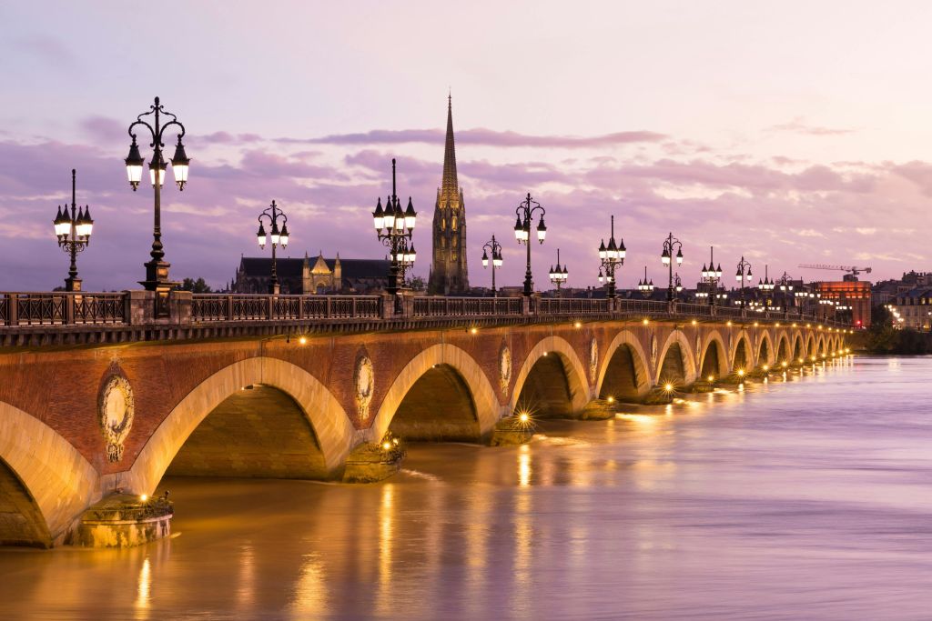 An illuminated stone bridge with lampposts spans a calm river at sunset, with a church spire visible in the background.