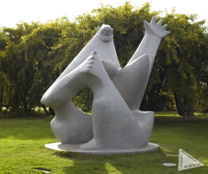 나무를 배경으로 잔디가 우거진 지역에 위치한 팔을 들어 올린 추상적인 흰색 조각상.