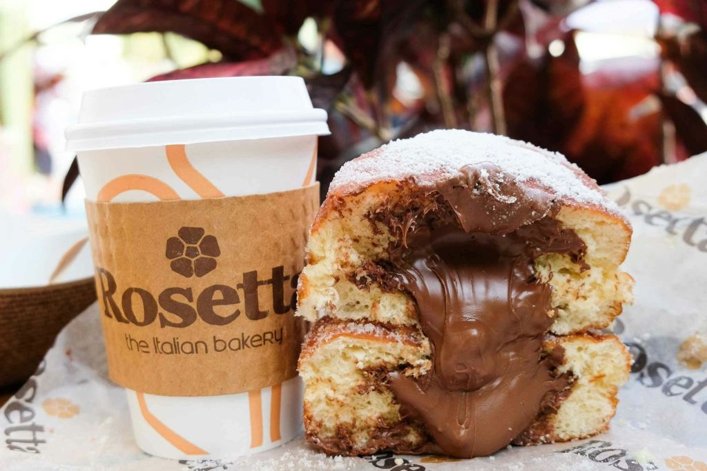 "이탈리아 빵집 로제타 (Rosetta the Italian bakery)" 라고 적힌 갈색 소매가 달린 커피잔 옆에 초콜릿이 떨어지는 크림이 가득한 페이스트리.