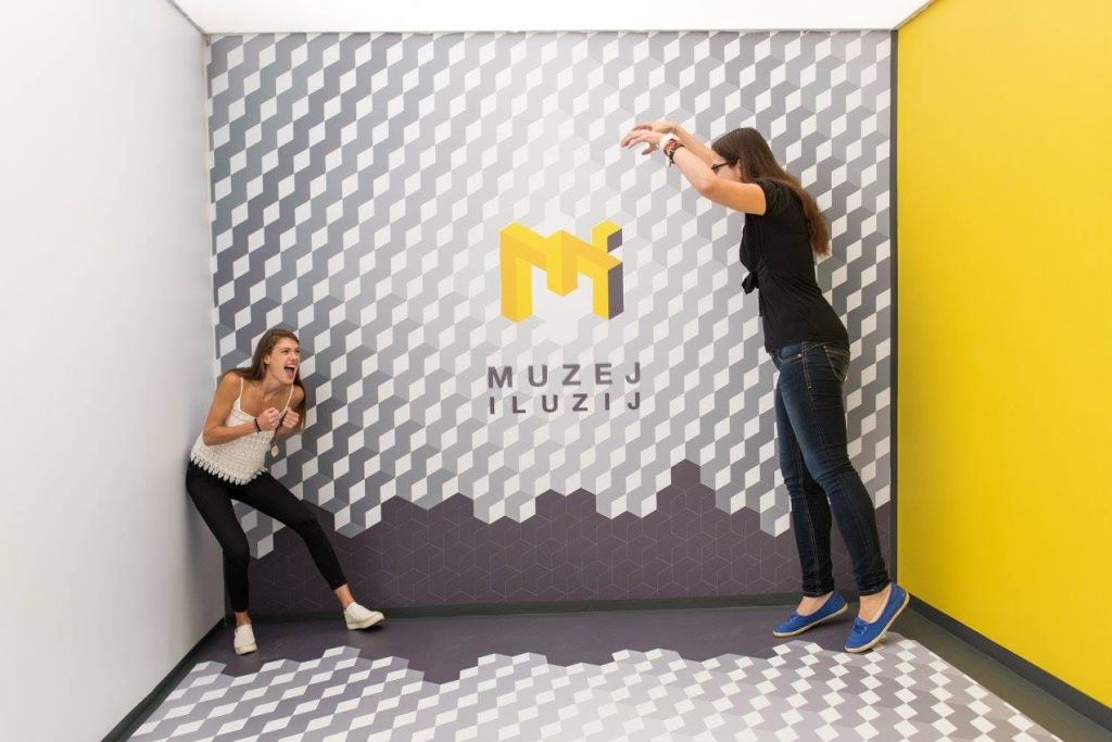 한 여성이 무늬가 있는 벽에 맞서고 있으며, 몸이 벗겨진 손이 아래로 뻗어 내려갈 때 무서워하는 것처럼 보입니다. 벽에는 "Muzej Iluzij" 라고 쓰여 있습니다.