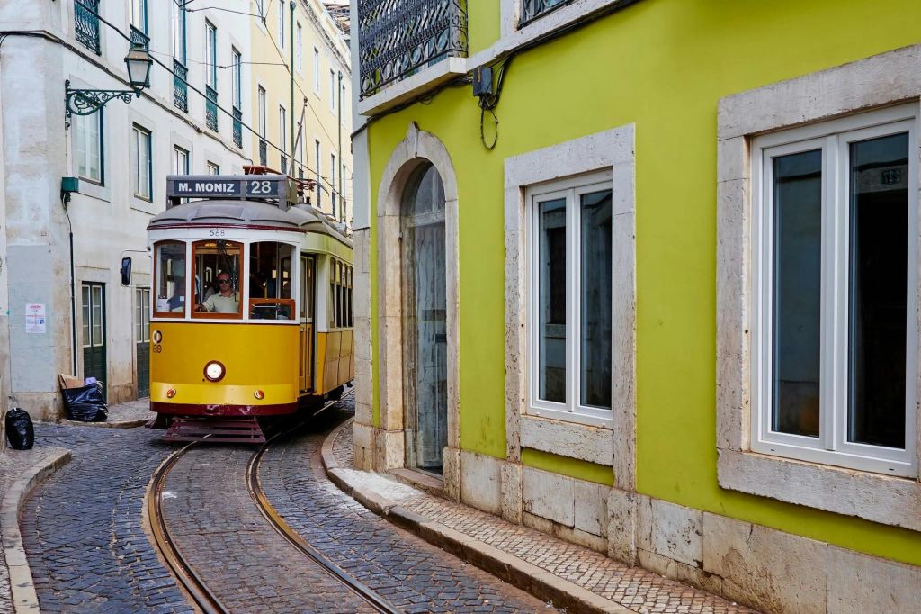 "28 M. Moniz" 라고 표시된 노란색 트램이 흰색으로 장식된 창문이 있는 녹색 건물 옆의 좁은 자갈길에서 굴곡을 이루고 있습니다.