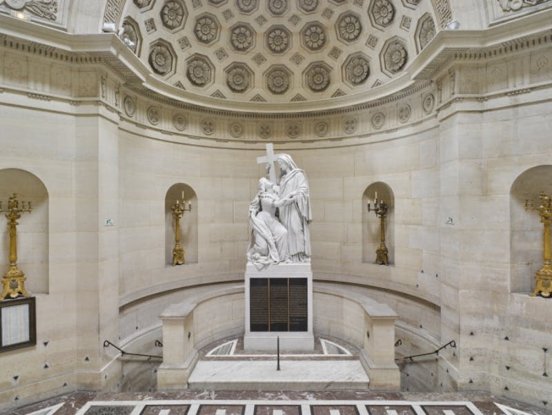 무릎을 꿇은 형상의 중앙 대리석 조각상과 십자가를 들고 서 있는 형상이 장식된 화려한 내부로, 두 개의 금색 촛대 옆으로 나란히 놓여 있습니다.