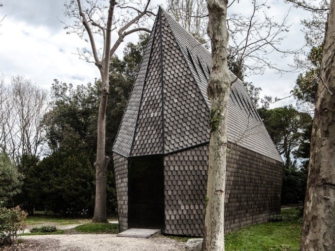 키가 큰 나무와 흙길이 이어지는 숲 속에 정강이 모양의 외관이 있는 작고 뾰족한 지붕 구조물이 서 있습니다.