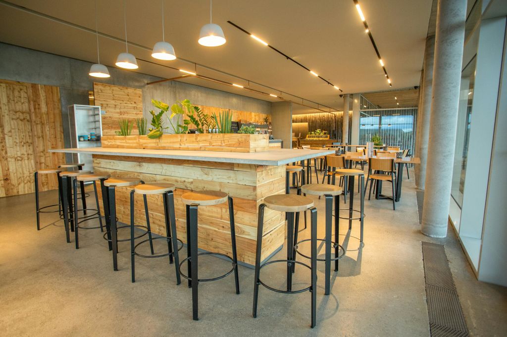 중앙 카운터에 나무 테이블, 높은 스툴, 매달린 램프, 실내 식물이 있는 현대적인 카페. 배경에 커다란 창문이 있습니다.